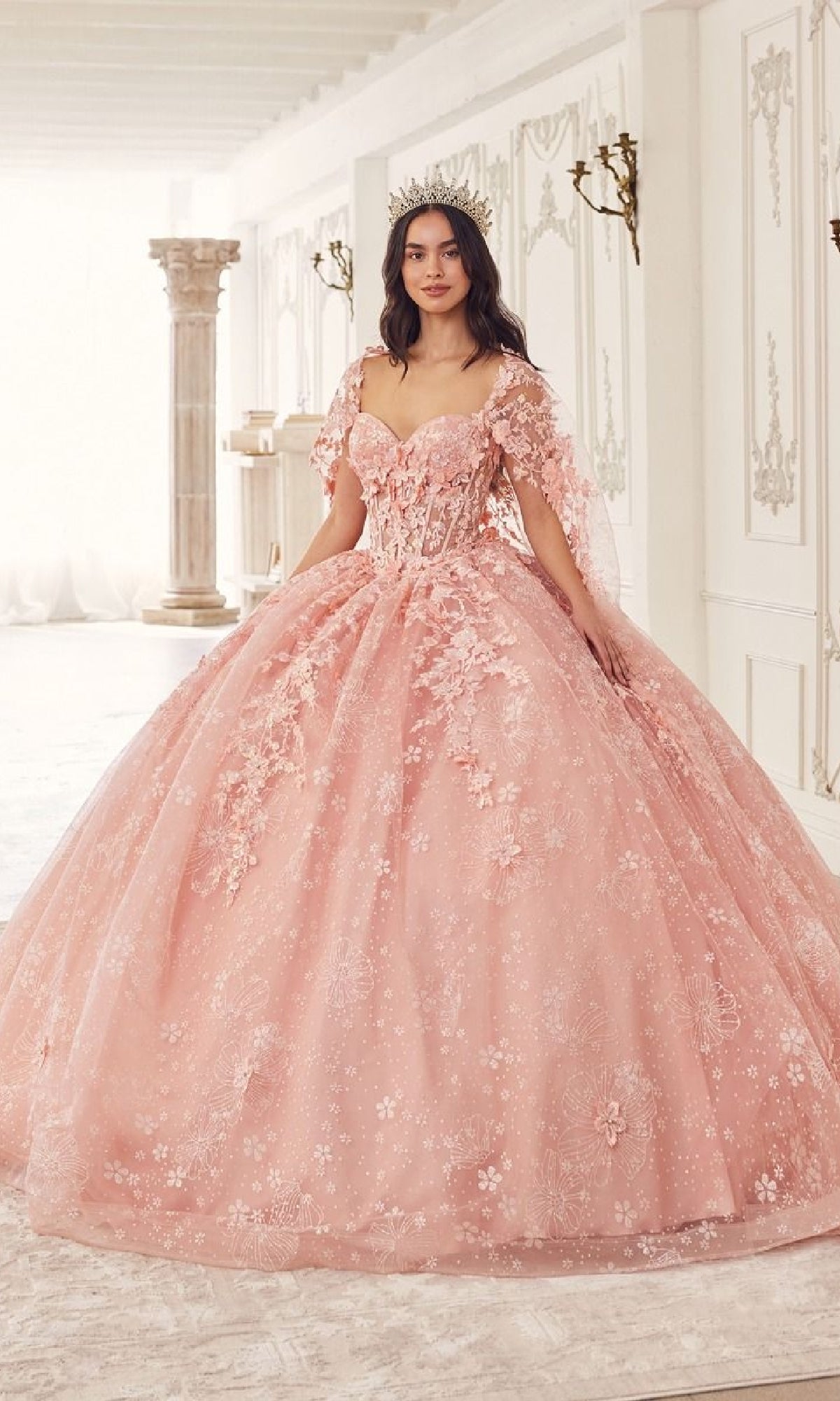 A woman stands indoors wearing the Quinceanera Dress 15719 by Ladivine—a pink, floral lace ball gown with a crown, set in a room with white walls and classical decor.